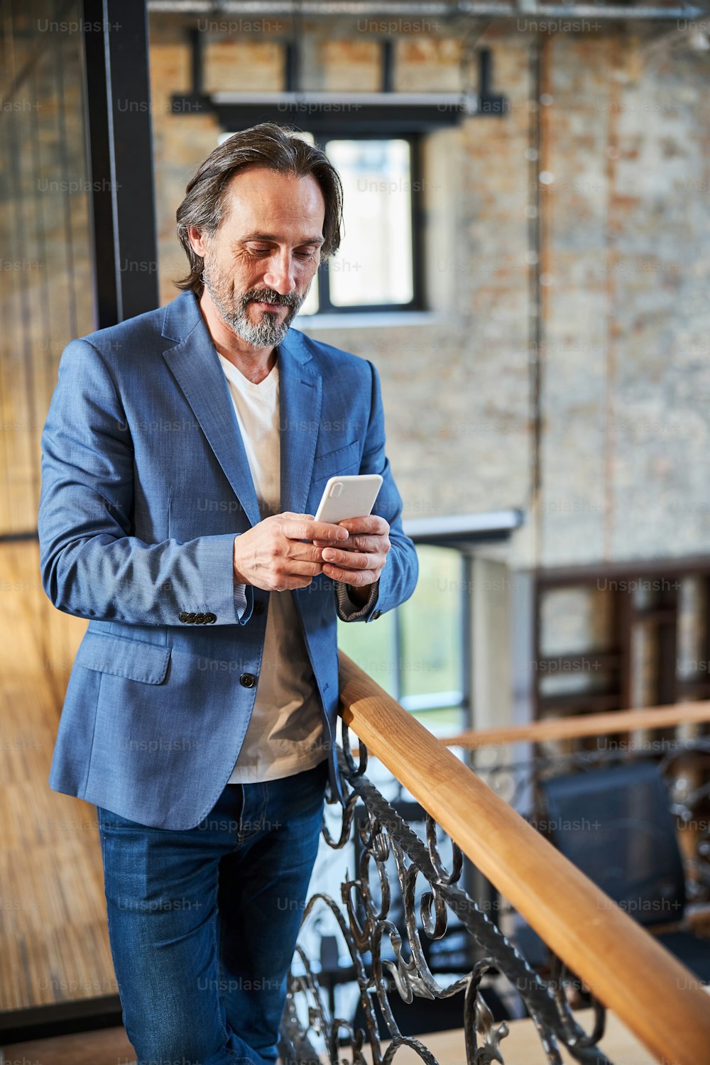 Carismático hombre barbudo con blazer azul mirando la pantalla de su teléfono inteligente mientras está parado cerca de la escalera