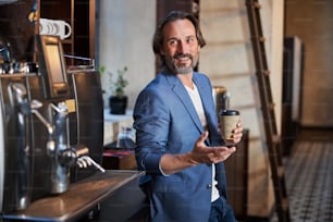 Waist-up photo of a joyful brunette man smiling while drinking coffee in a barista corner and gesturing