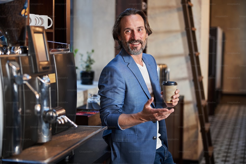 Foto dalla vita in su di un gioioso uomo bruno che sorride mentre beve un caffè in un angolo barista e gesticola