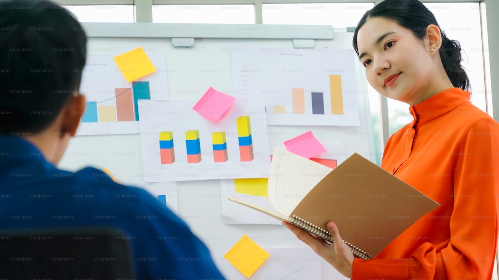 Young woman explains business data on white board in casual office room . The confident Asian businesswoman reports information progress of a business project to partner to determine market strategy .