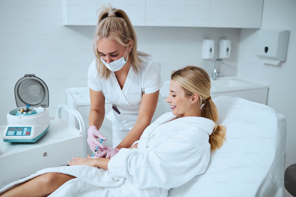 Cheerful blonde Caucasian female patient undergoing the hand rejuvenation treatment in a modern beauty salon