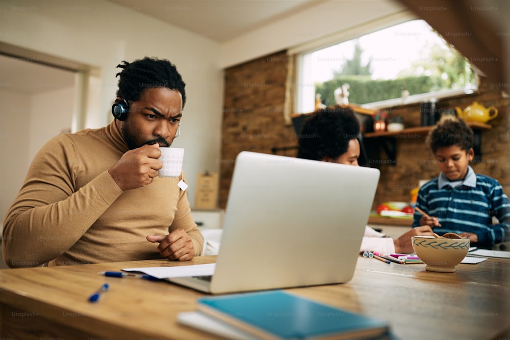 Black working father using laptop while having cup of tea at home. Mother and son are in the background.