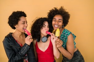 Retrato de un grupo multiétnico de amigos que se divierten y disfrutan del verano mientras comen helado sobre fondo amarillo.