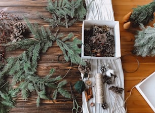 Making christmas rustic wreaths with pinecones, scissors,thread and berries on wooden rustic table top view. Festive home decor. Happy holidays and Merry Christmas!