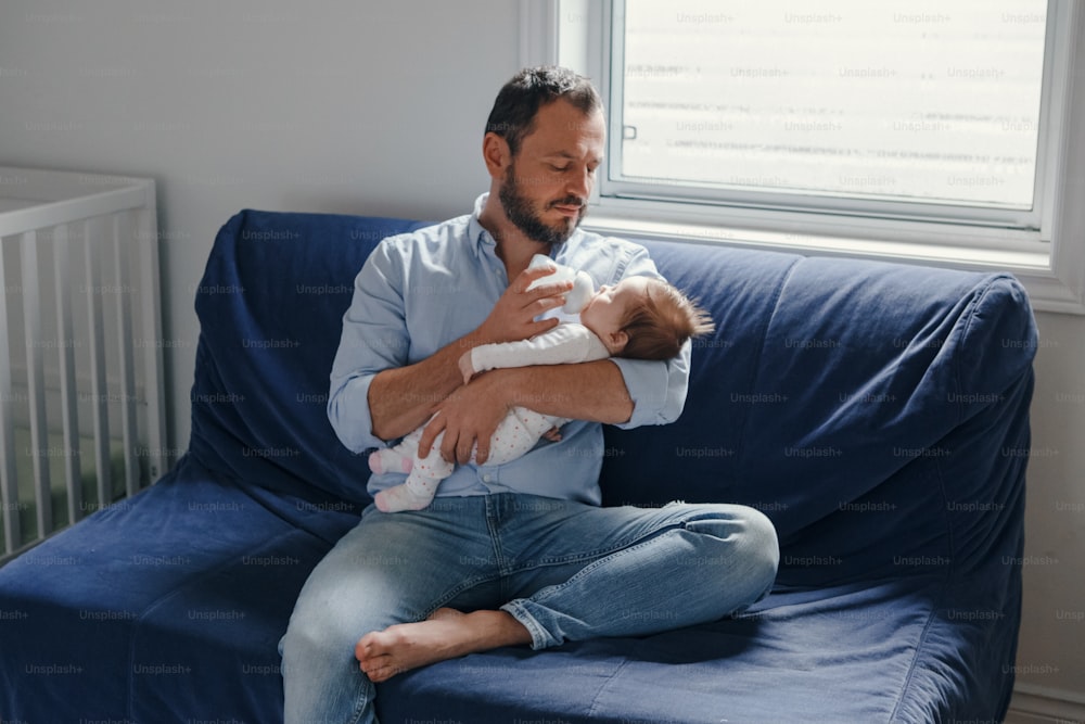 Middle age Caucasian father feeding newborn baby from bottle milk. Man parent holding rocking child on his hands. Authentic lifestyle candid real moment. Single dad family home life concept.