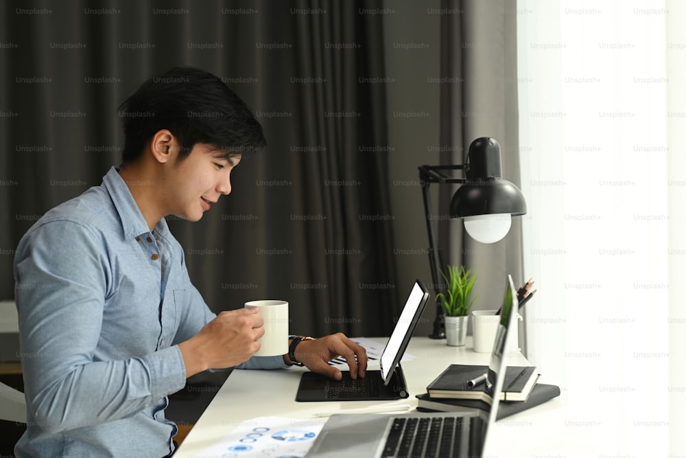 Vista lateral de un diseñador gráfico está trabajando con una tableta de computadora y tomando café en su espacio de trabajo por la mañana.