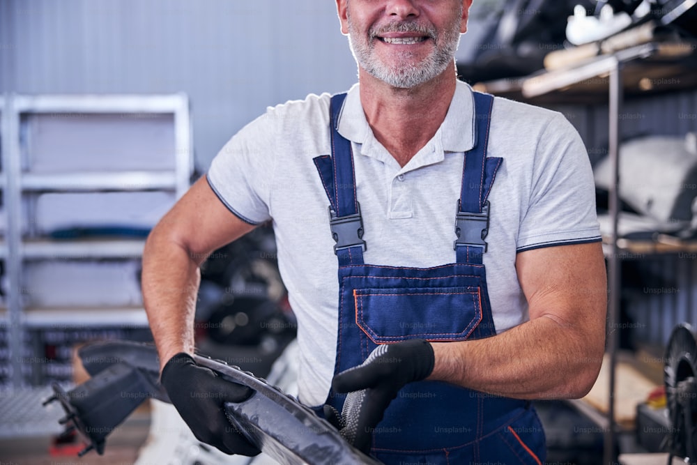 Trabajador masculino barbudo alegre sosteniendo la parte del automóvil y sonriendo mientras trabaja en el garaje
