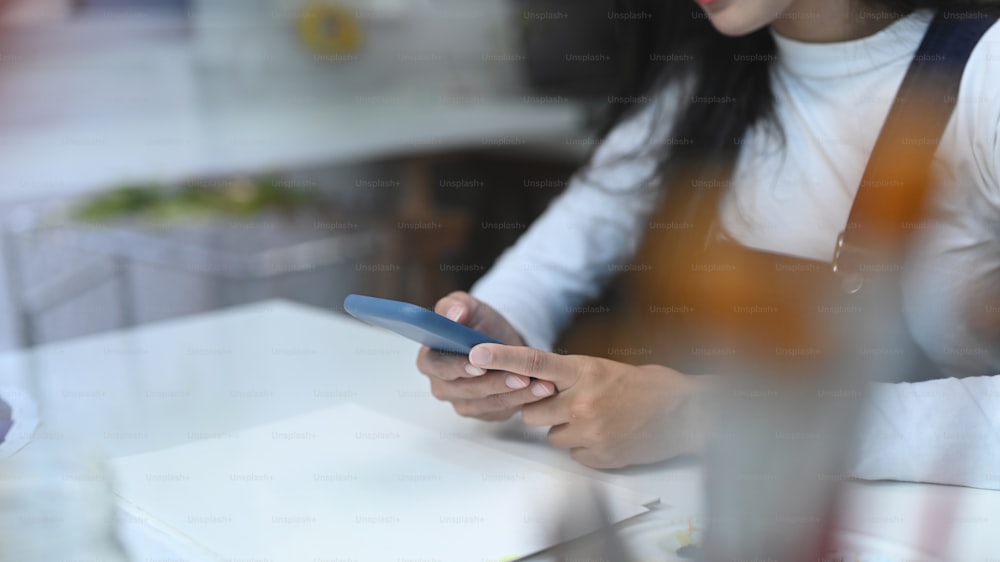 Cropped shoto der jungen Frau, die am Schreibtisch sitzt und auf den Smartphone-Bildschirm schaut, surft drahtloses Internet auf Gadget.