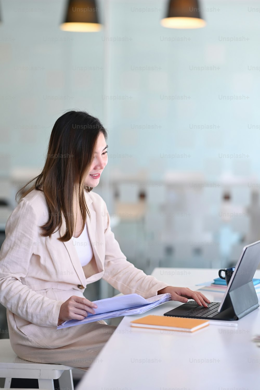 Una mujer de negocios concentrada escribiendo en una computadora portátil y sosteniendo papeles preparando un informe que analiza los resultados del trabajo en la oficina.