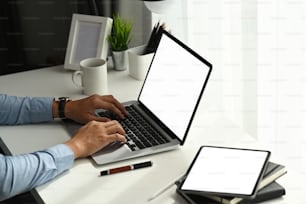 Young man hands working on laptop at comfortable workspace with blank screen tablet on desk.