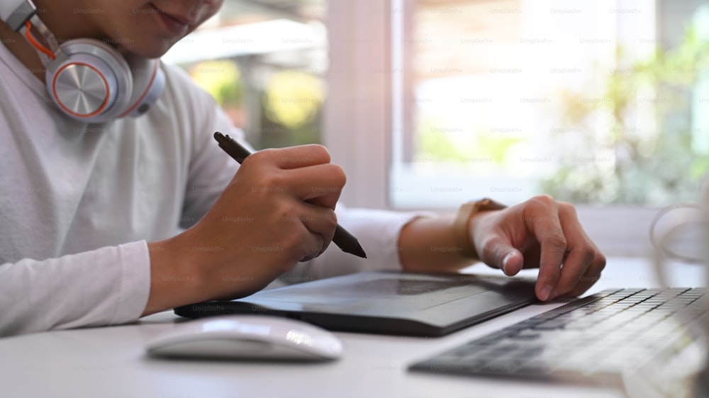 Foto cortada do designer criativo masculino usando tablet sem fio moderno com teclado e caneta stylus enquanto trabalha no escritório moderno.