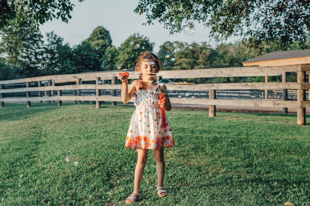 Ritratto candido di bambina caucasica in età prescolare che soffia bolle di sapone nel parco al tramonto estivo. Vero autentico momento felice dell'infanzia. Stile di vita bambini attività stagionale all'aperto.