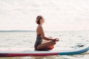 Young Caucasian woman practising yoga on paddle sup surfboard at sunset. Female stretching doing workout on lake water. Modern individual hipster outdoor seasonal summer sport activity.