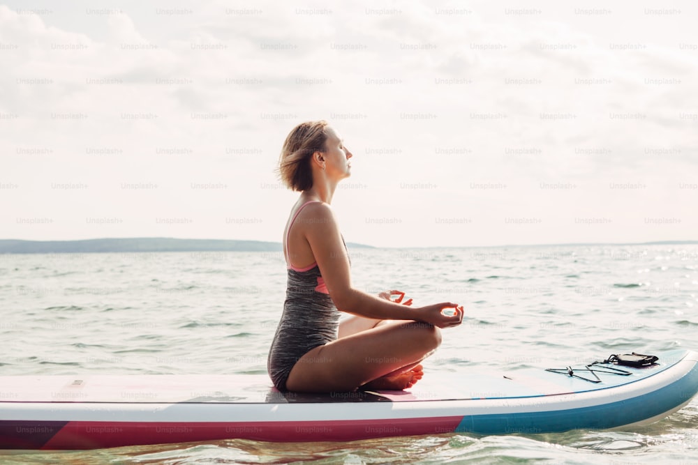 Giovane donna caucasica che pratica yoga sulla tavola da surf sup paddle al tramonto. Stretching femminile che fa allenamento sull'acqua del lago. Moderna attività sportiva estiva stagionale all'aperto hipster individuale.