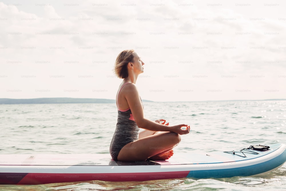 Young Caucasian woman practising yoga on paddle sup surfboard at sunset. Female stretching doing workout on lake water. Modern individual hipster outdoor seasonal summer sport activity.