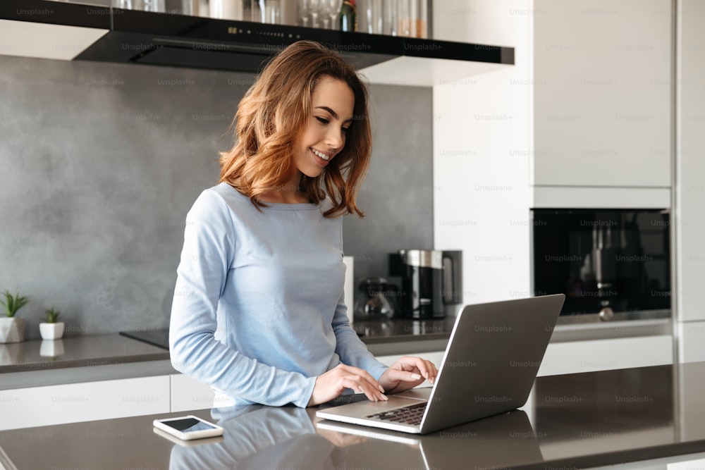 Portrait of a joyful young woman using laptop computer while standing at the kitchen table at home