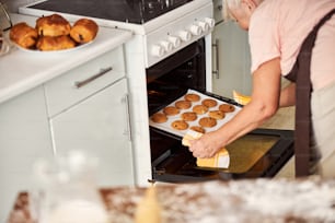 Foto recortada de una mujer de cabello gris inclinada hacia adelante y sacando una sartén caliente de galletas frescas del horno
