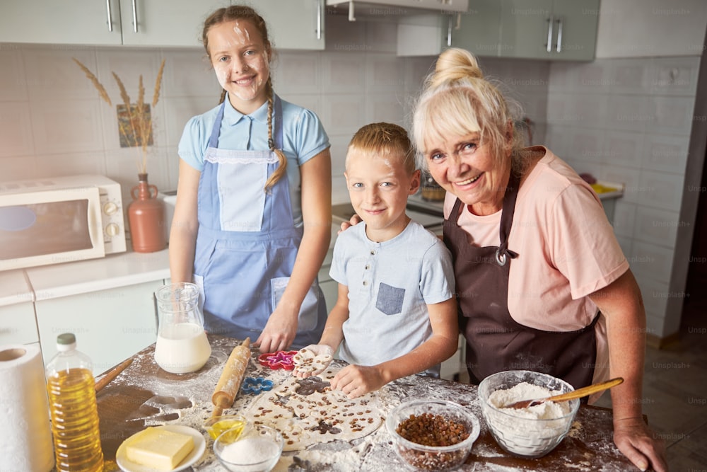 Delightful elderly lady posing with her lovely grandchildren near kitchen table