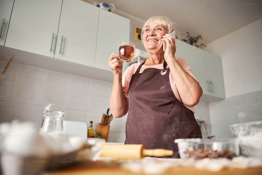 Signora anziana contenta che sorride mentre beve il tè e parla al telefono nella sua cucina