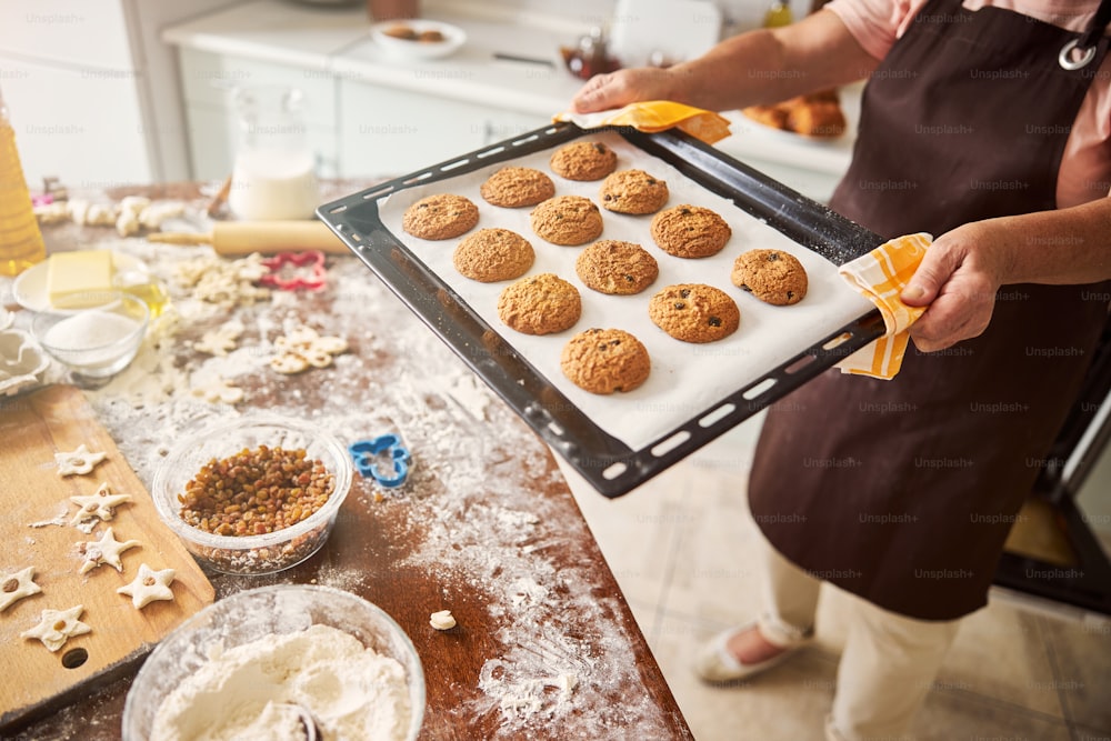 Cropped photo of woman in apron using towels to carry hot metal tray with freshly-baked load of cookies