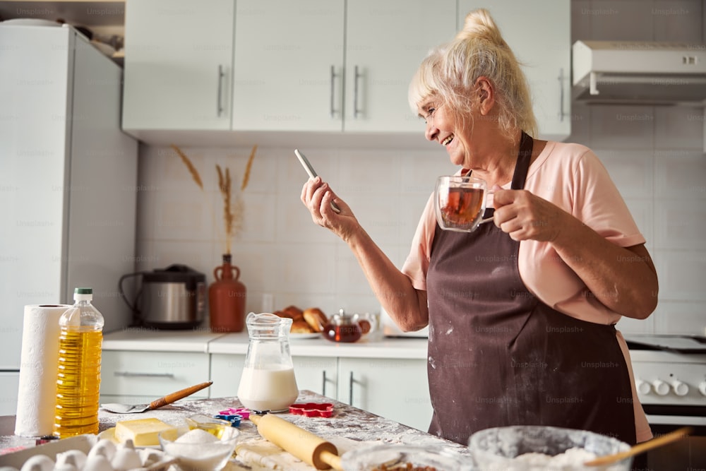 Excited aged lady laughing and looking at her smartphone while drinking tea in kitchen