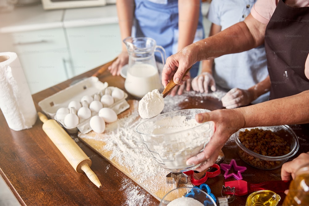 Cropped photo of senior woman holding wooden spoon full of flour while showing her grandchildren how to make dough