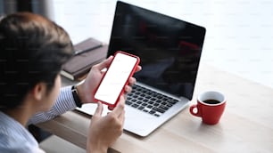 Cropped shot of male using mobile phone with blank screen and sitting in front of laptop in office.