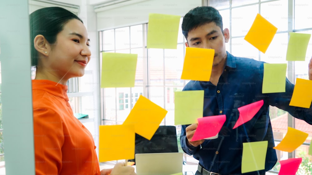 Business people work on project planning board in office and having conversation with coworker friend to analyze project development . They use sticky notes posted on glass wall to make it organized .