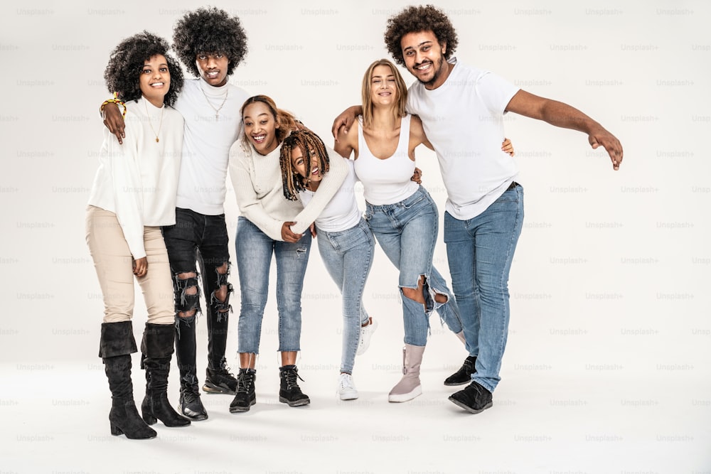 カメラを見ながら、白いシャツとジーンズを着て、白いスタジオの背景に一緒にポーズをとっている幸せな若い友人の多民族グループの写真。ミレニアル世代の友情ライフスタイルのコンセプト。