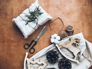 Christmas stylish eco gift flat lay, eco friendly furoshiki present with scissors, pine cones and festive decorations on wooden rustic table. Happy holidays and Merry Christmas!