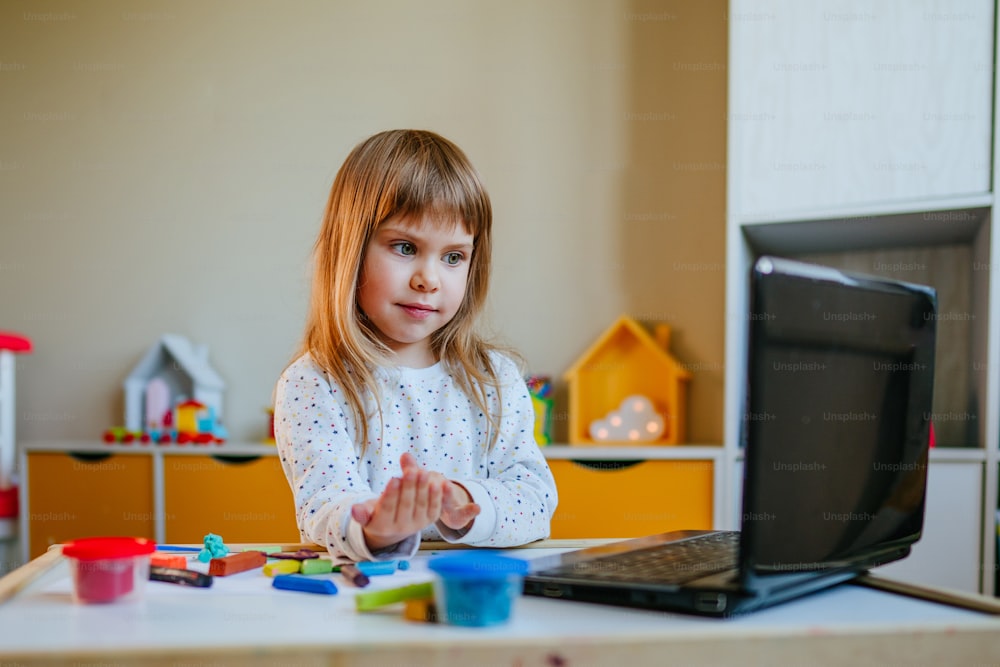 어린 소녀는 실내에서 노트북으로 온라인 학습 수업을 보고 다채로운 점토를 성형합니다. 원격 가정 학습 개념입니다.