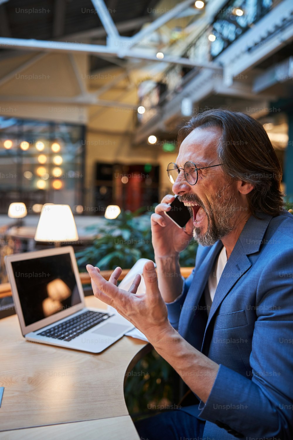 Shocked brunette man yelling with surprise while talking on the phone and gesturing wildly at a co-working space