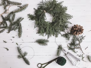 Making christmas rustic wreath, cedar and fir branches with scissors on white wooden rustic table, top view. Festive simple handmade home decor. Happy holidays and Merry Christmas!