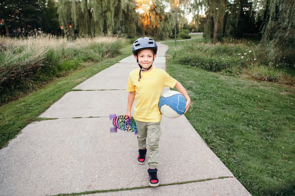 Happy Caucasian boy in helmet holding ball and skateboard in park on summer day. Seasonal outdoor children activity sport. Healthy childhood lifestyle. Kids individual summer sport.