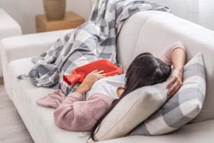 Dor de estômago aliviada por termóforo enquanto a mulher se deita em um sofá sob o cobertor.