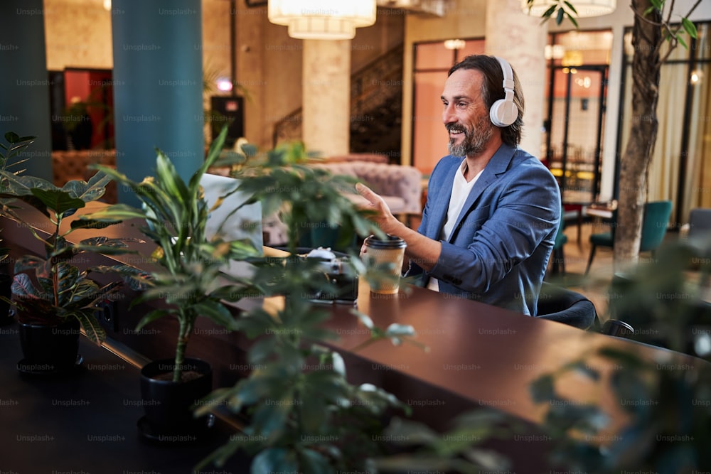 Vivacious middle-aged man in headphones smiling and gesturing in front of laptop screen while lounging in hotel lobby