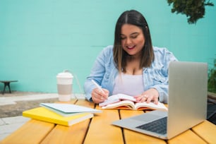 Portrait d’une jeune femme de grande taille étudiant avec un ordinateur portable et des livres tout en étant assise à l’extérieur dans un café.