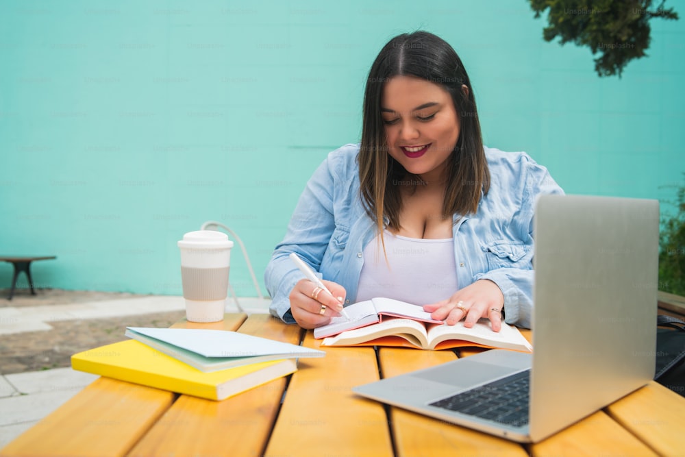 Retrato da jovem mulher plus size que estuda com laptop e livros enquanto está sentada ao ar livre no café.