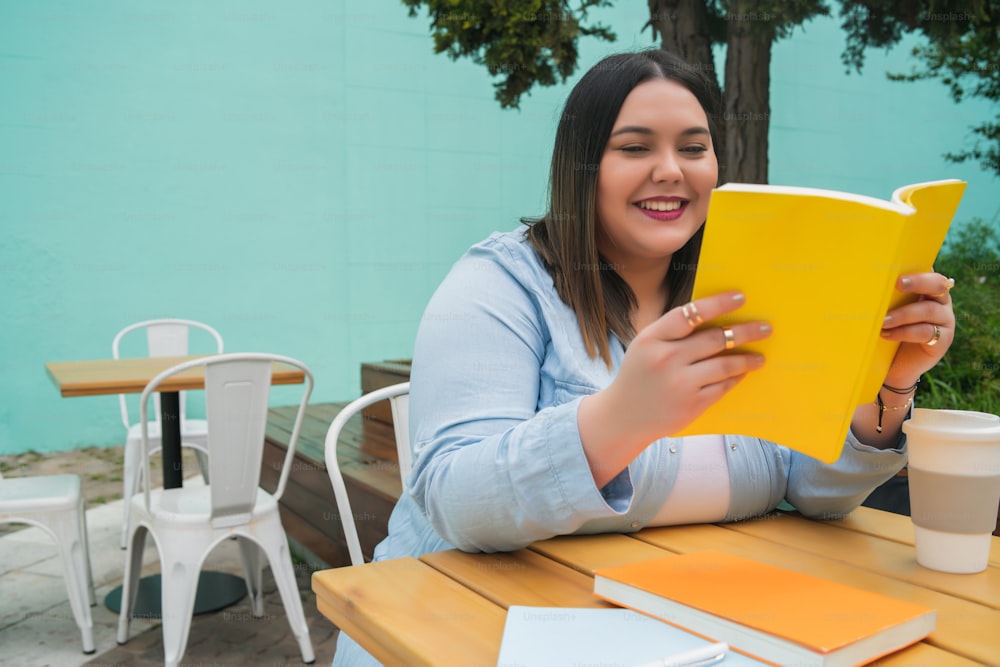 Portrait of young plus size woman enjoying free time and reading a book while sitting outdoors at coffee shop.