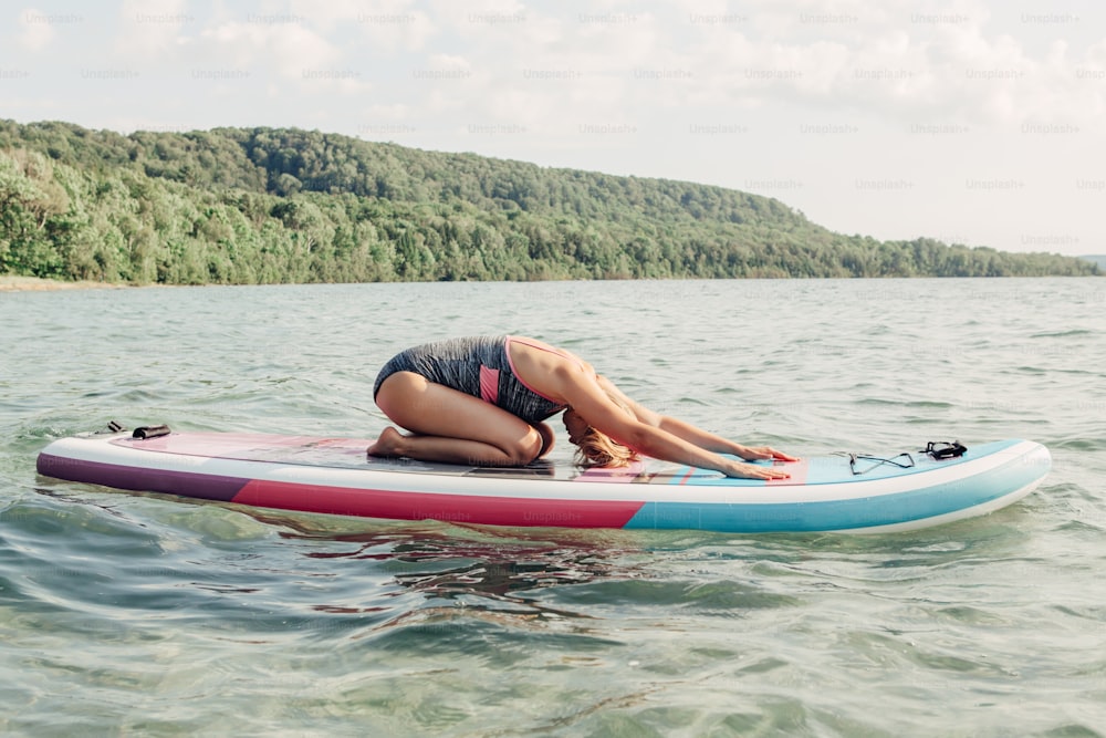 Young Caucasian woman practising yoga on paddle sup surfboard at sunset. Female stretching doing workout on lake water. Modern individual hipster outdoor seasonal summer sport activity.