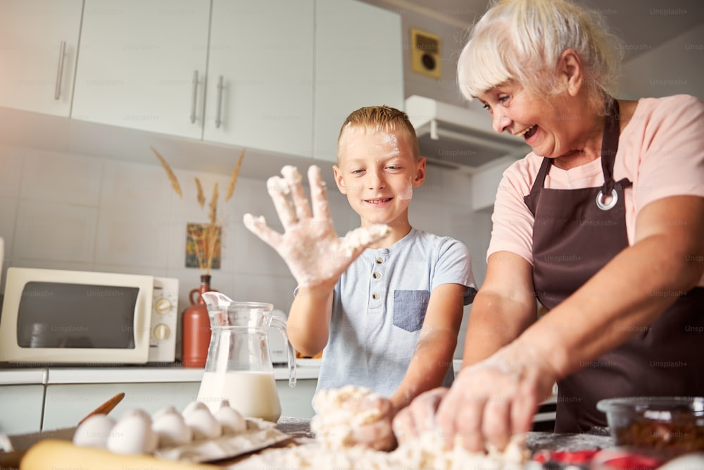 Amicable elderly lady looking at joyous boy having his hand covered in dough while cooking at the table