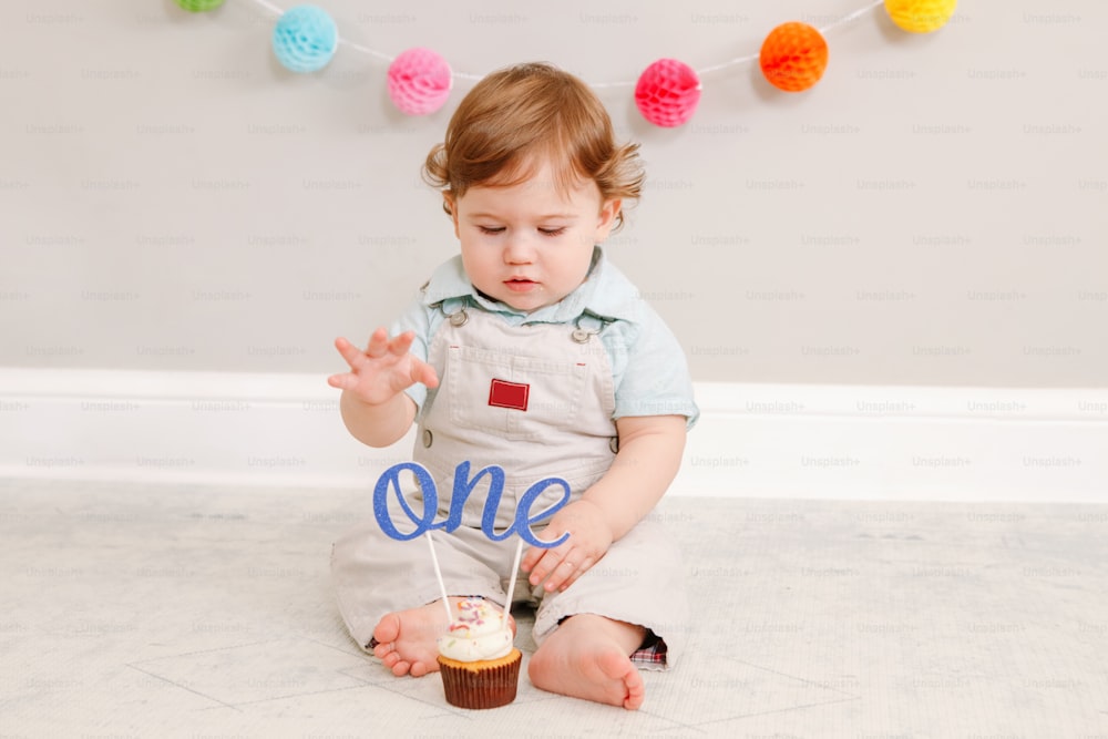 Happy cute Caucasian baby boy celebrating his first birthday at home. Child kid toddler sitting on floor in room. Tasty cupcake dessert with cake topper word one. Happy birthday concept.