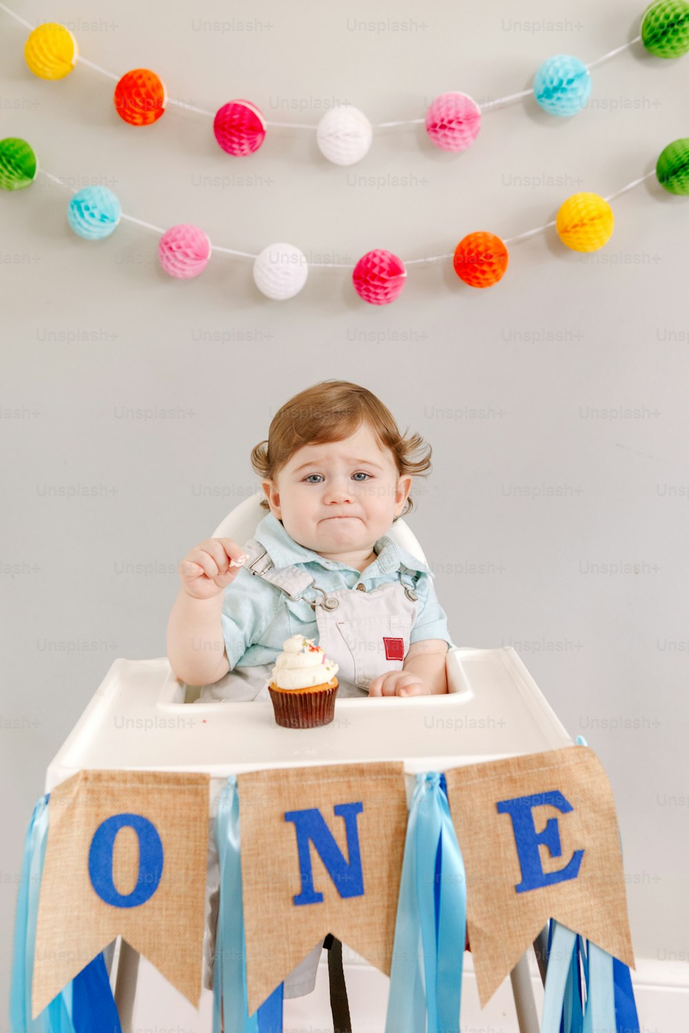 Cute adorable sad upset Caucasian baby boy celebrating his first birthday at home. Pensive child kid toddler sitting in high chair table eating tasty cupcake dessert. Happy birthday lifestyle concept.