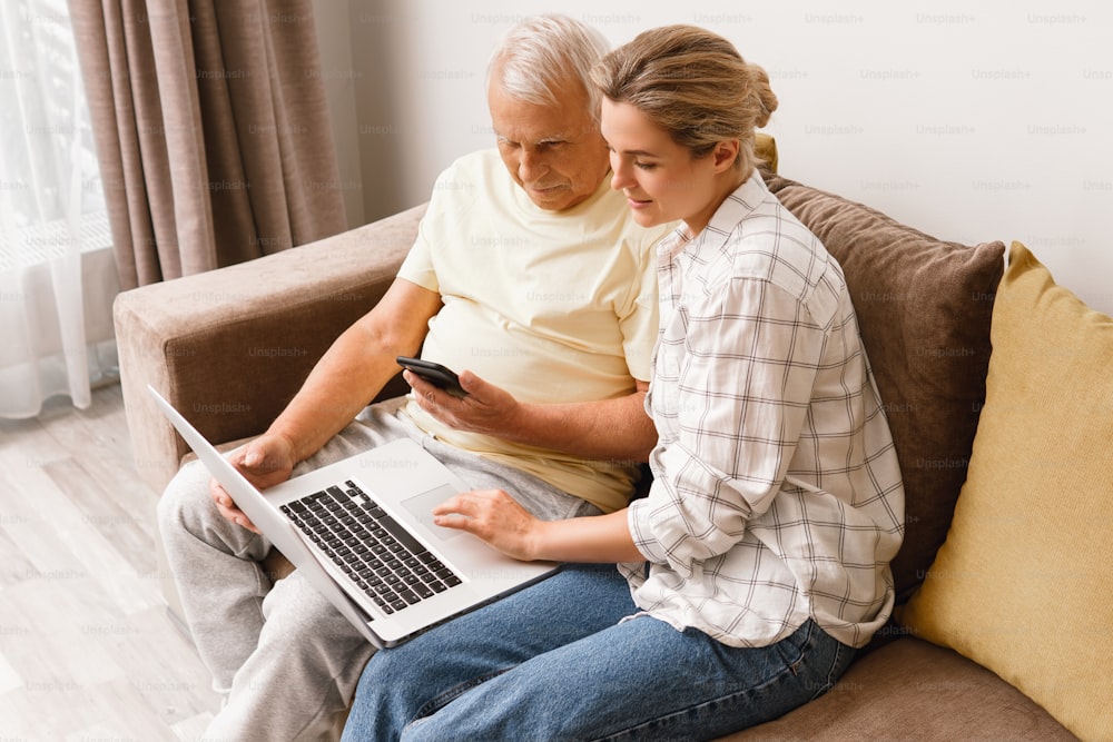 Young woman explaining to elderly man how to use laptop and smatphone