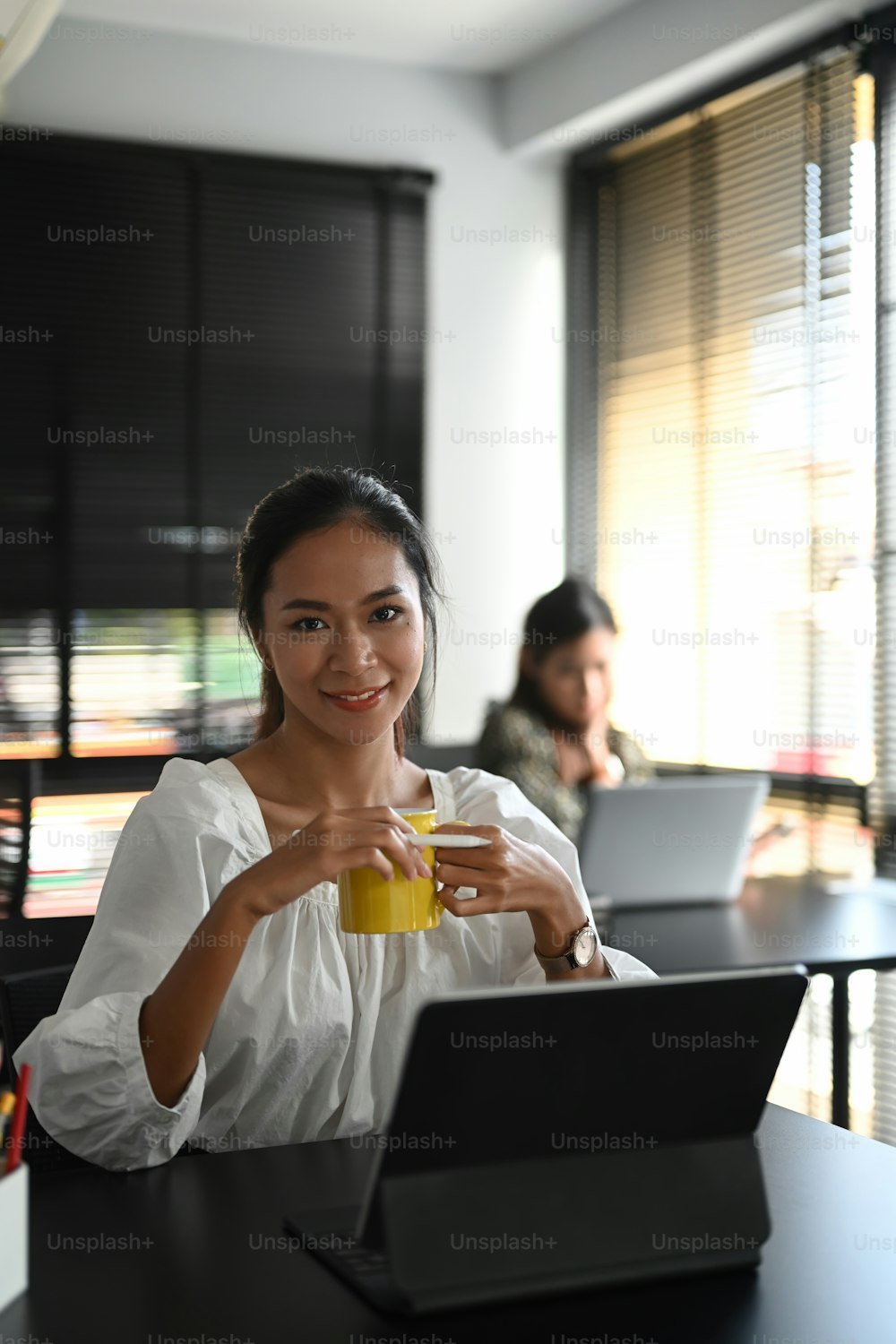 Portrait of business woman smiles at camera while holding coffee cup and her colleague sitting in the background.
