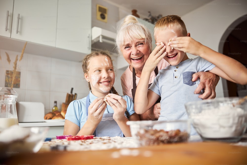 Alegre anciana y sus nietos riendo y jugando con masa para galletas mientras posan cerca de la mesa de la cocina desordenada