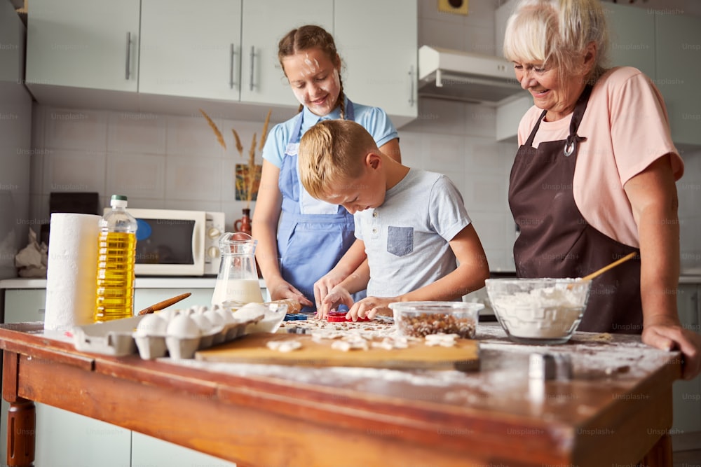 Konzentrierter blonder Junge, der Teig zu einem Keks formt, während seine Schwester und Oma ihn beobachten