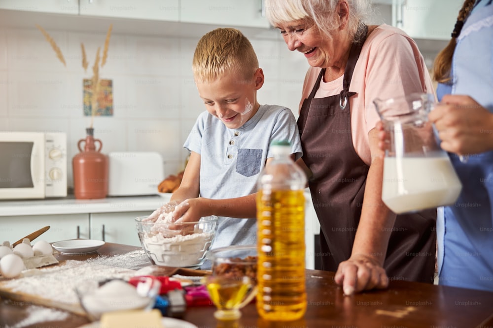 Cheerful grandma looking at contented boy with a handful of flour while cooking in kitchen with her grandchildren