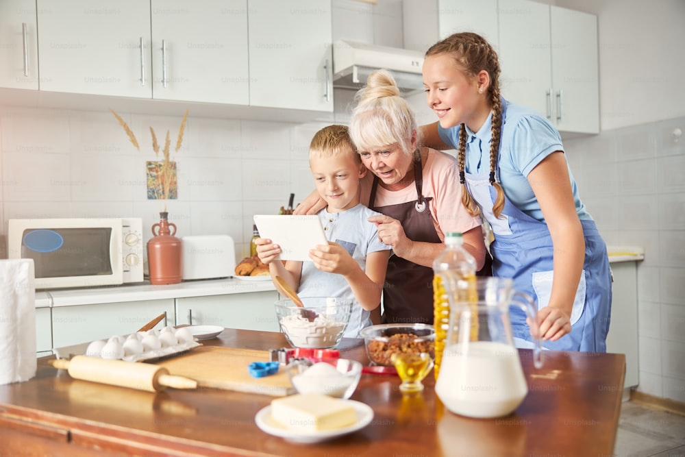 Enthusiastic senior lady and two children looking at tablet screen while standing by the kitchen table