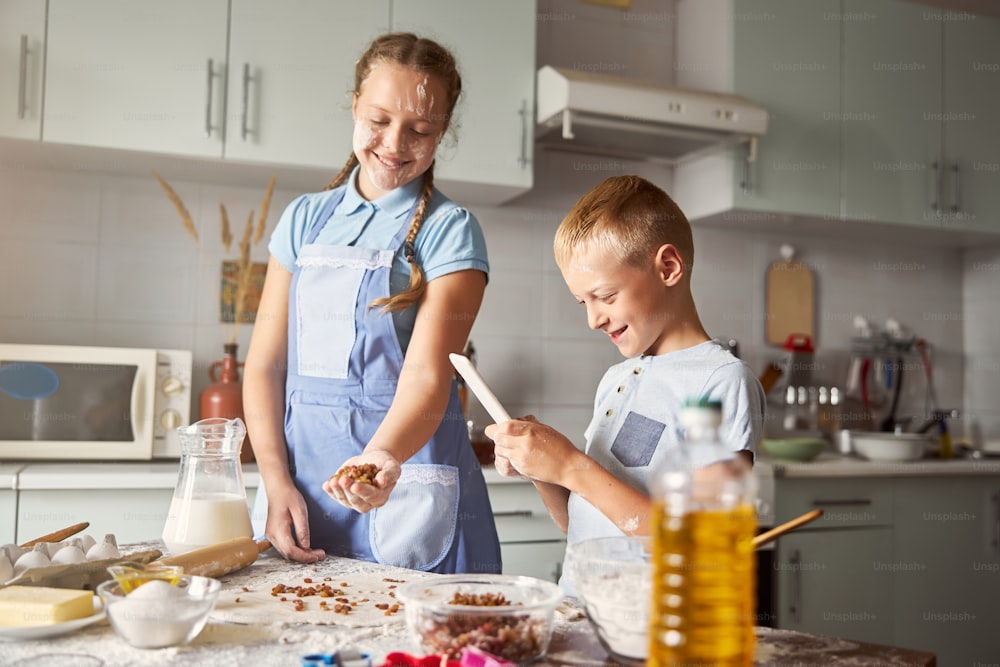 Glad teenage girl smiling and holding handful of raisins while her younger brother looking at his smartphone